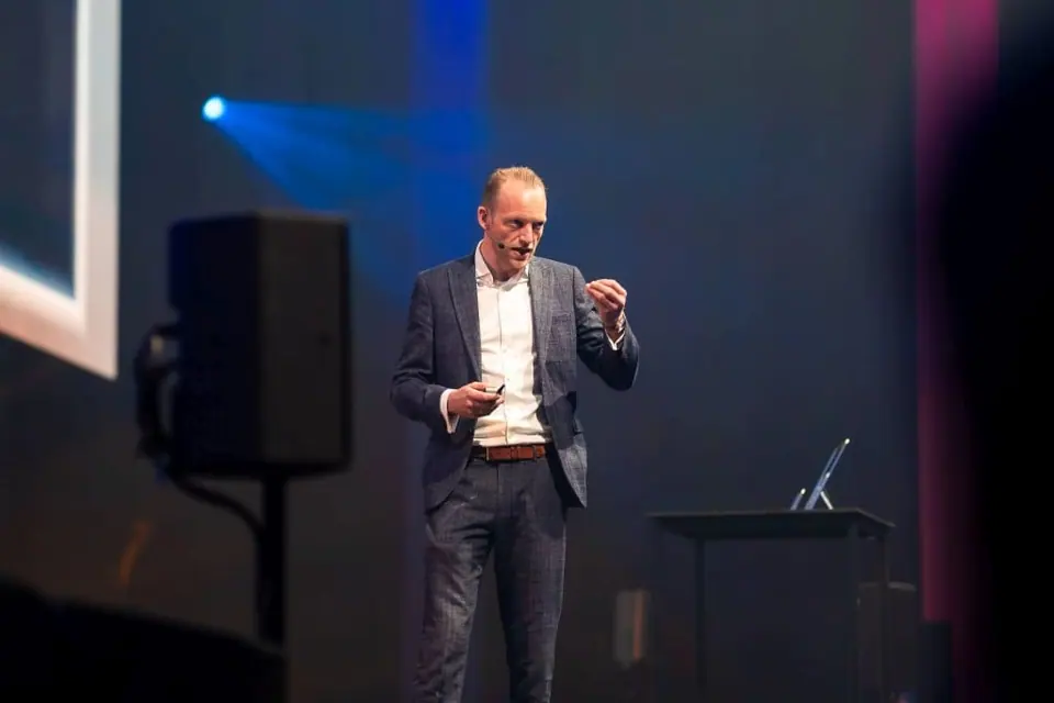 Stef Verbeeck presenting on a stage lit in blue, next to a loudspeaker