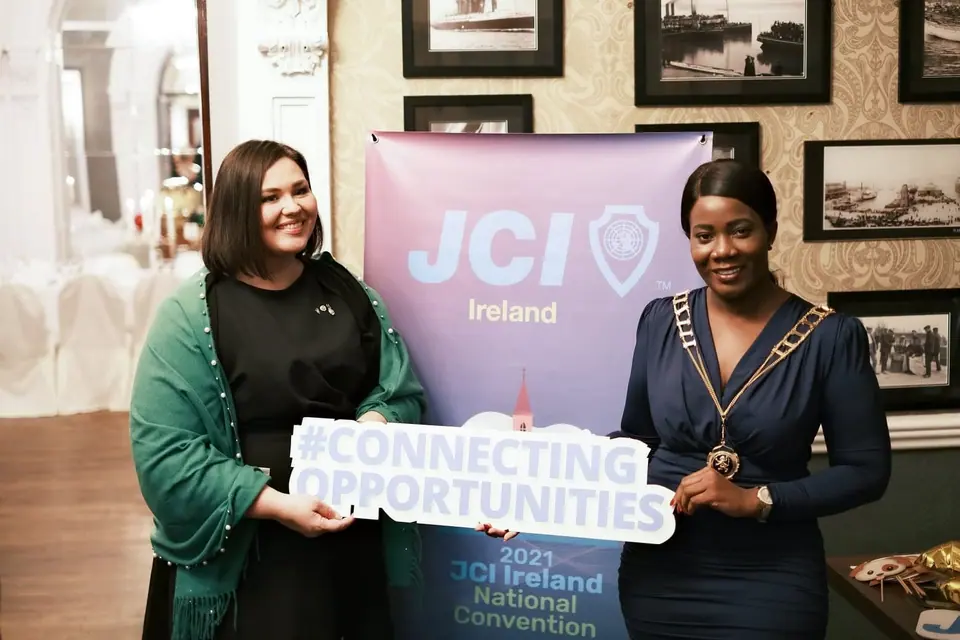 Two women at a JCI Ireland National Convention, one wearing a presidential chain of office, holding a Connecting Opportunities sign