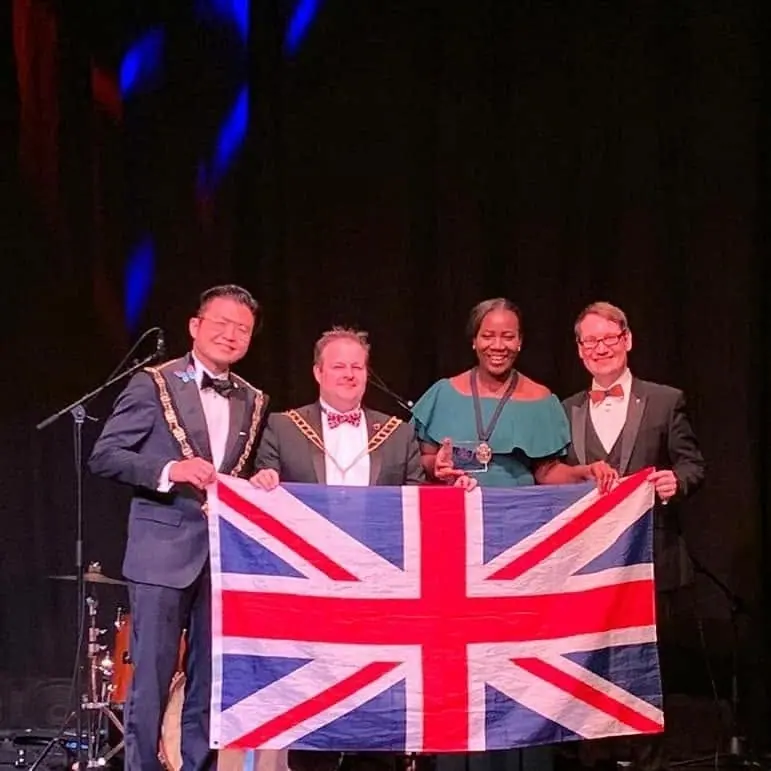 Four people in formal wear on stage holding a Union Jack flag, two of them wearing chains of office