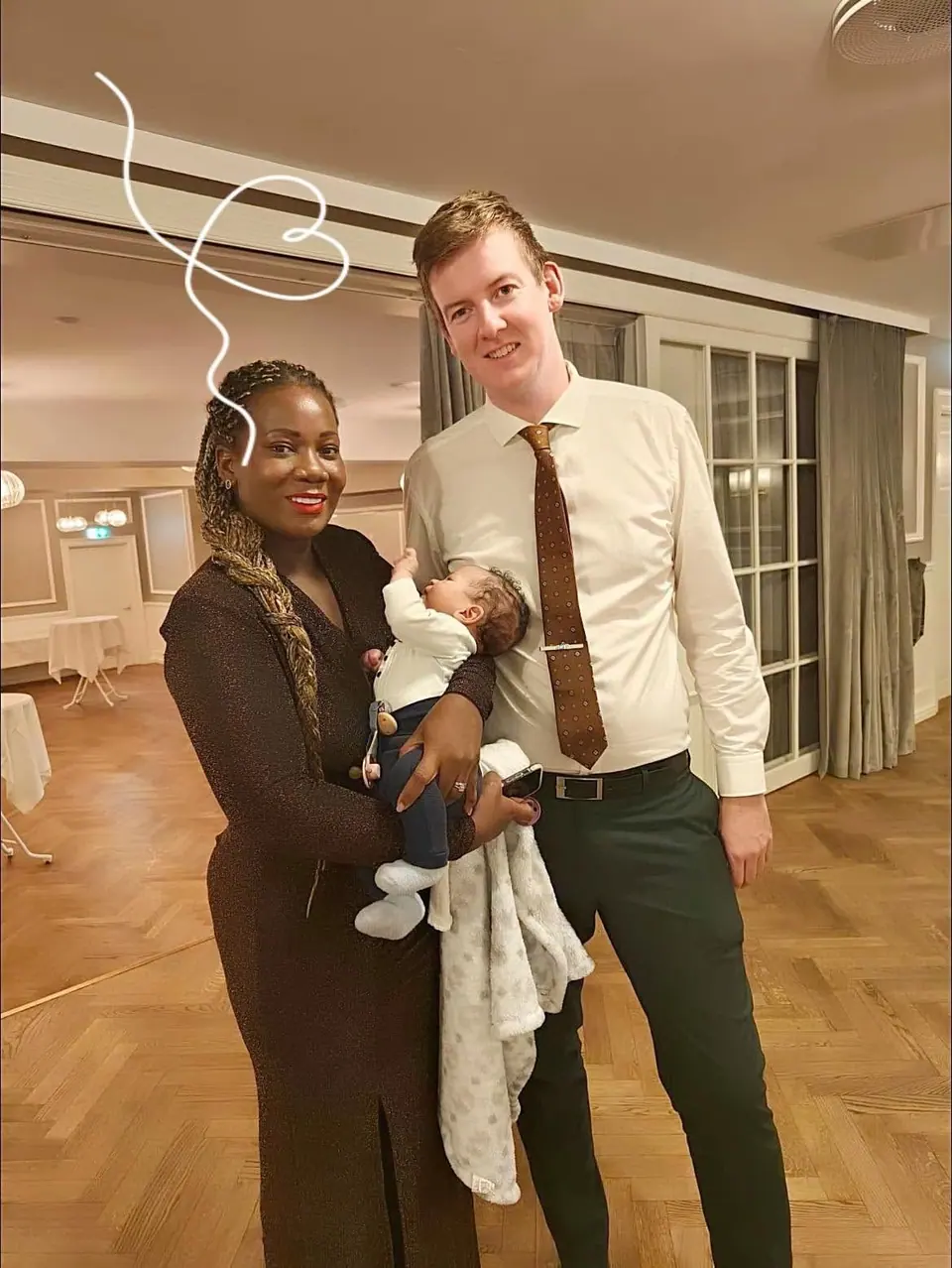A woman holding a baby standing beside a man in a shirt and tie in a function room