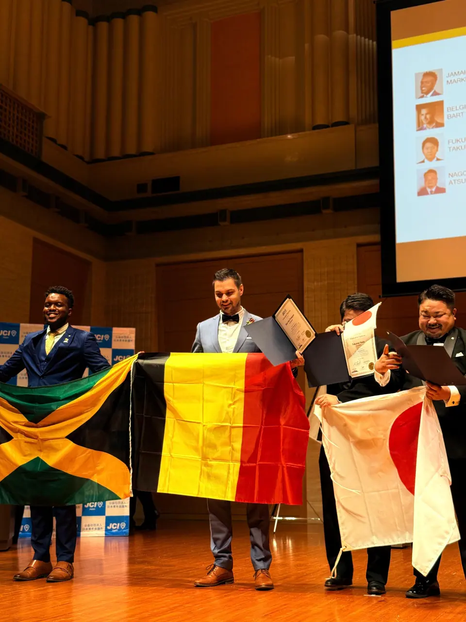 Three men on a concert-hall stage holding Jamaican, Belgian and Japanese flags