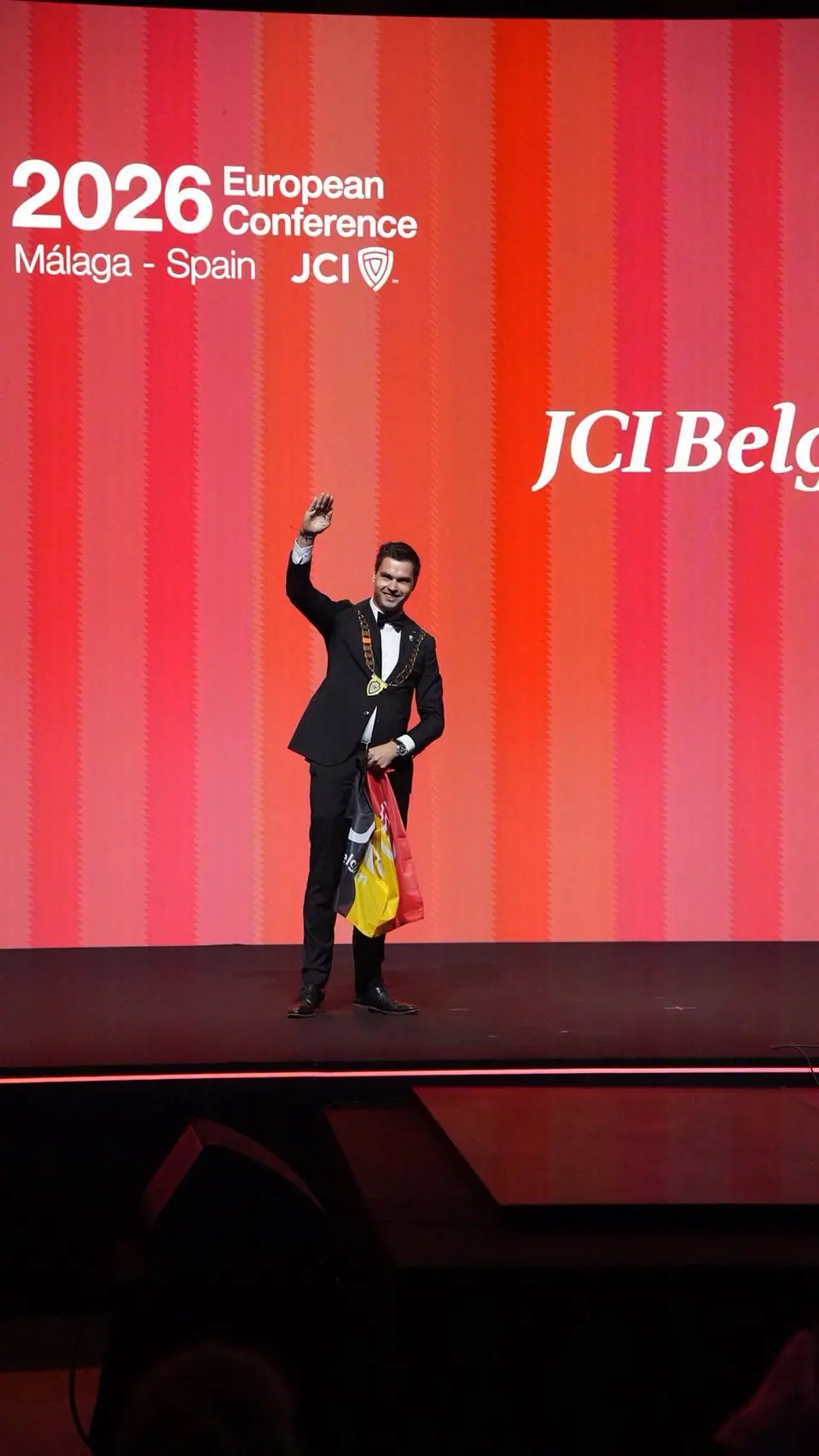 A man on stage waving a flag under a JCI Belgium and 2026 European Conference Málaga screen