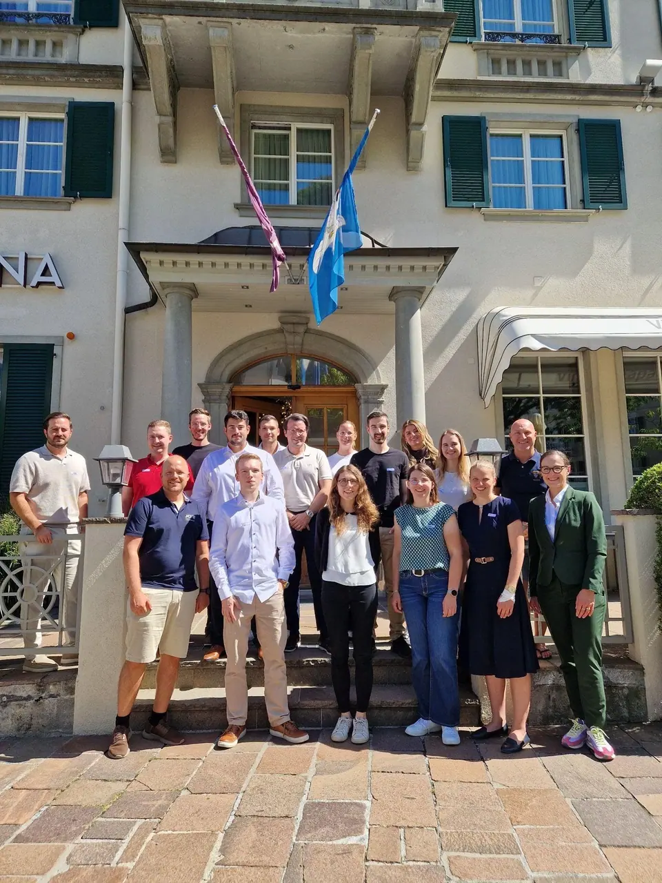 A group photo of about twenty people in front of a hotel with a JCI flag