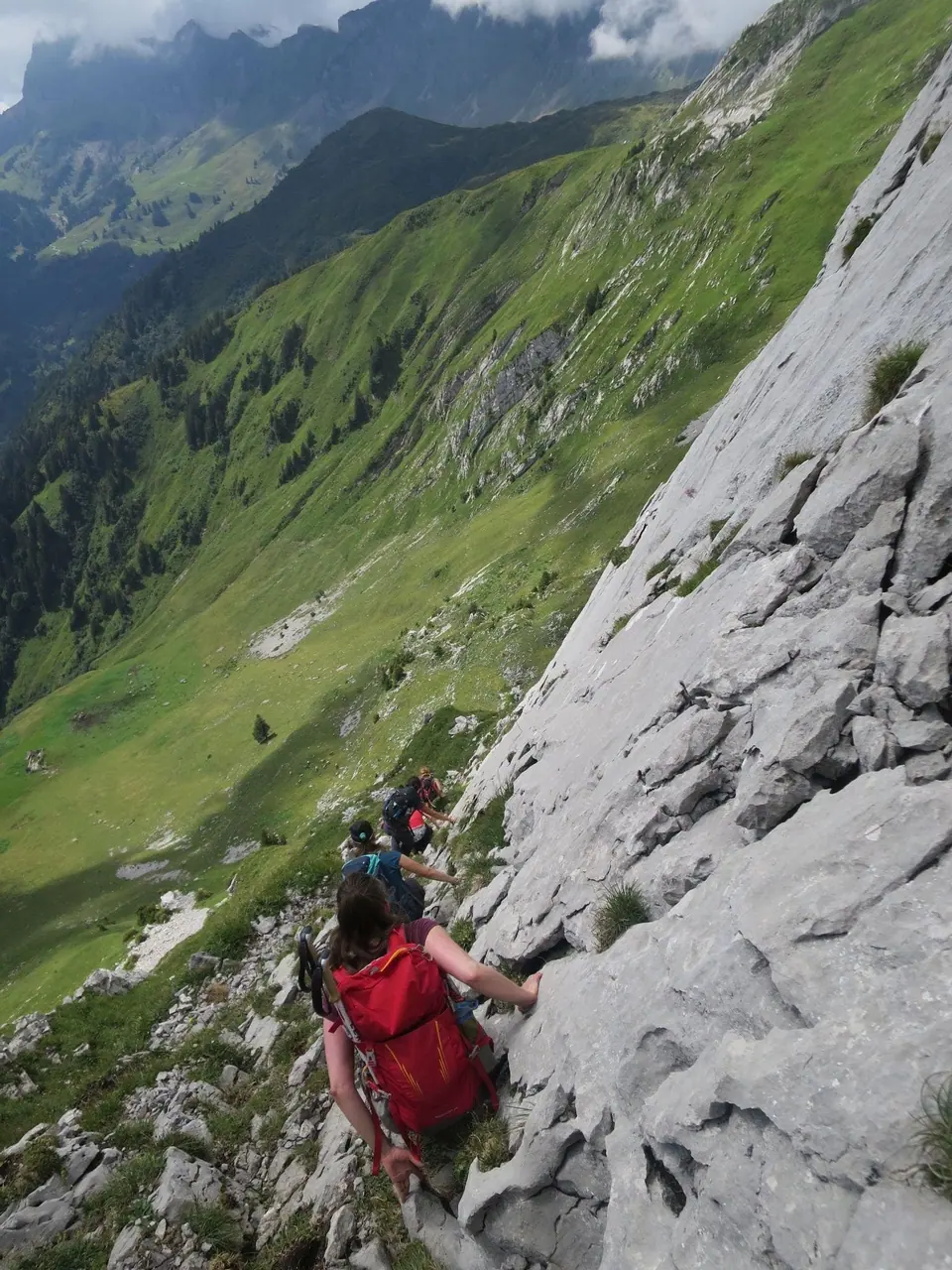 A mountain guide in a red jacket climbing a rocky slope above a green alpine valley
