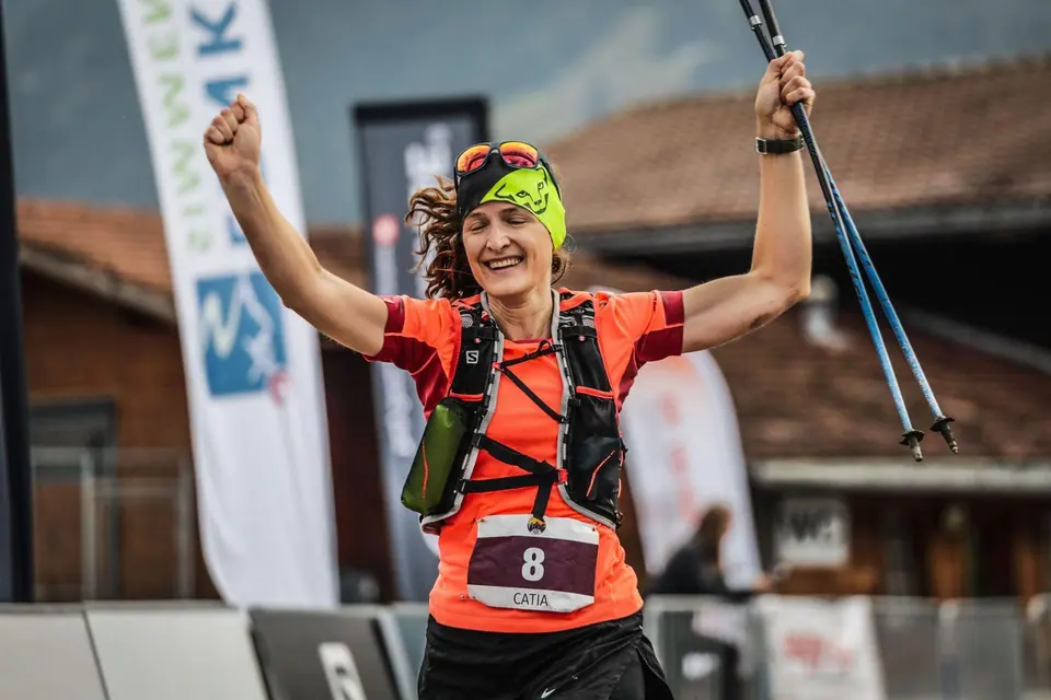 A trail runner with raised arms and poles crossing the finish of a mountain race, bib number 8