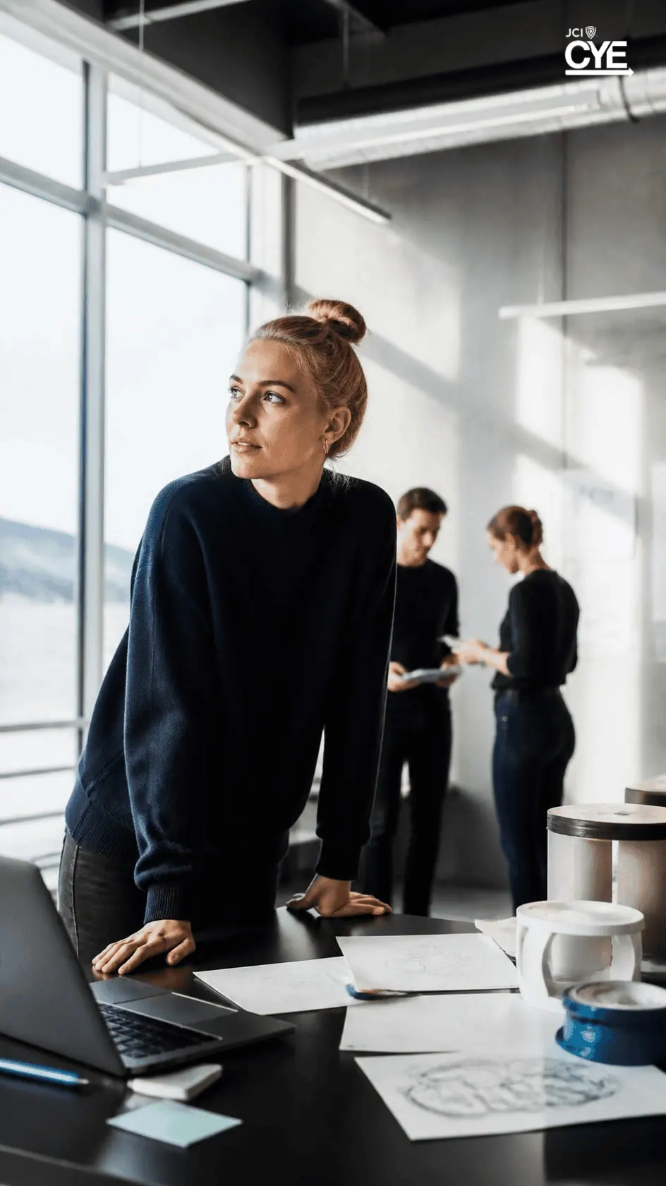 A founder leaning over a desk with a laptop and papers while colleagues talk behind her